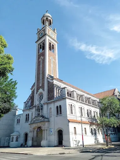 El templo visto desde la esquina: campanario de ladrillo con reloj y fachada blanca bajo el cielo celeste